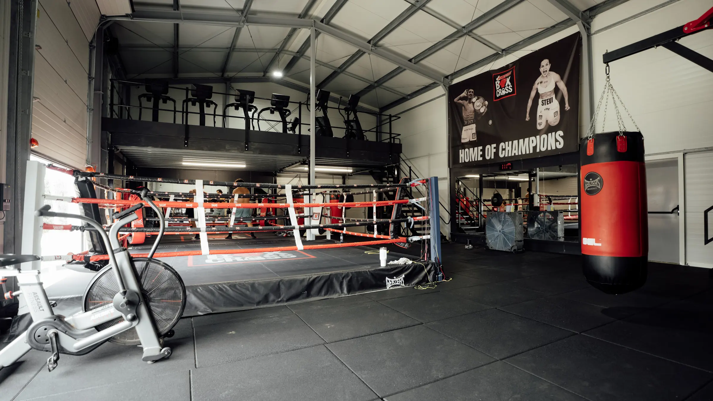 The full-size competition boxing ring at BoxCross UK seen from the mezzanine, with heavy bags and the Home of Champions banner
