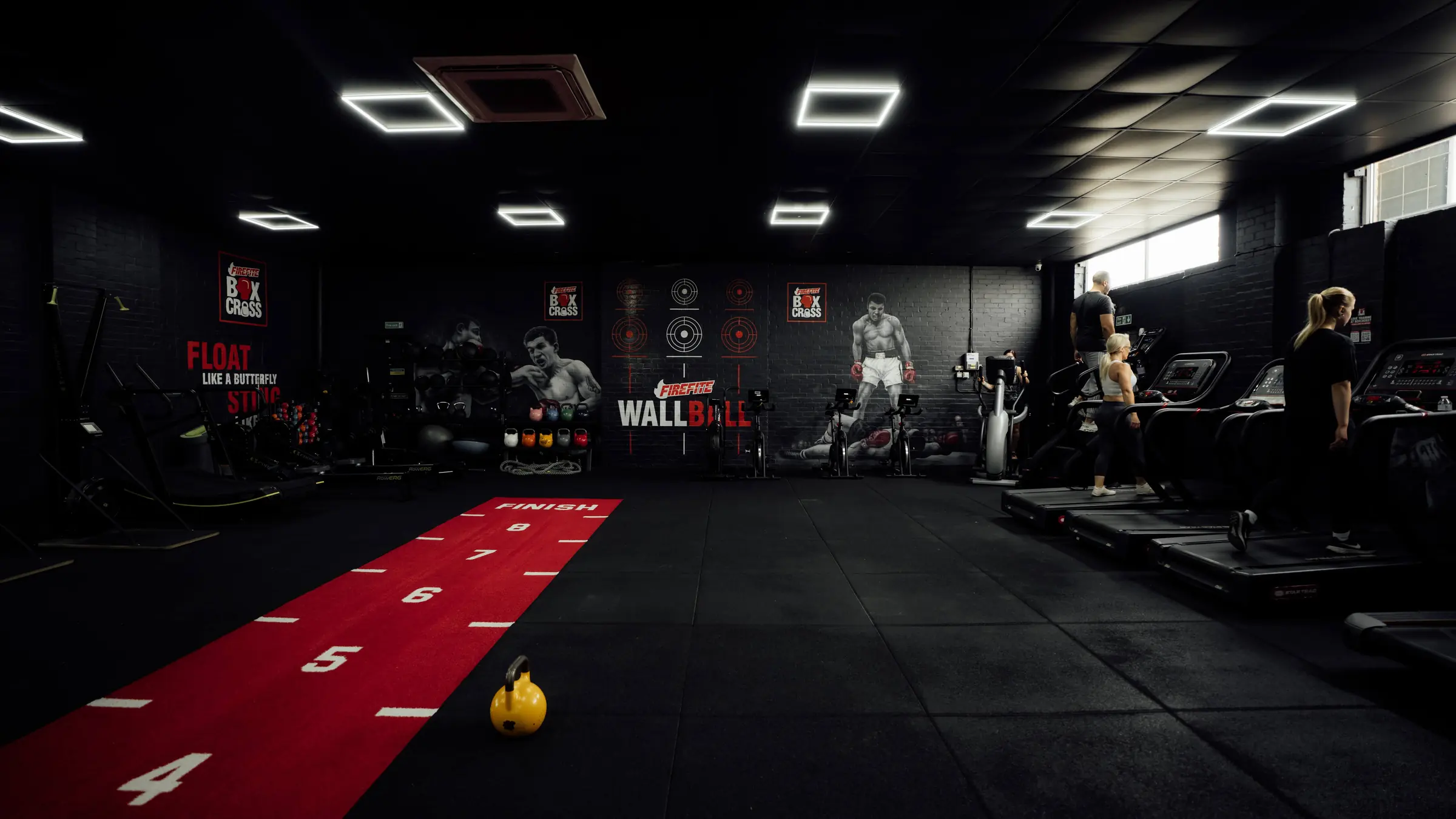 The functional room at BoxCross UK, a 24 hour gym in Wisbech: the red sled track running towards the wall ball wall, with members on the treadmills under the window