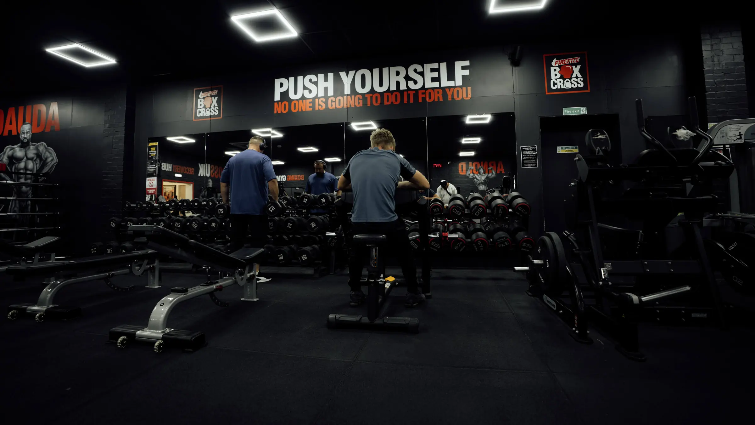 Two members picking dumbbells off the rack under the Push Yourself sign on the main floor at BoxCross UK Wisbech