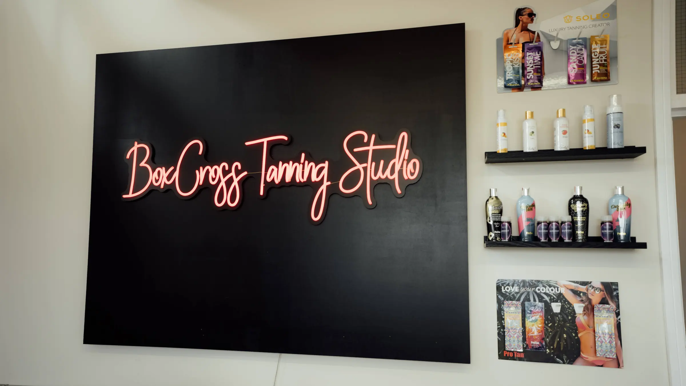 The BoxCross Tanning Studio neon sign beside shelves of tanning creams and accelerators